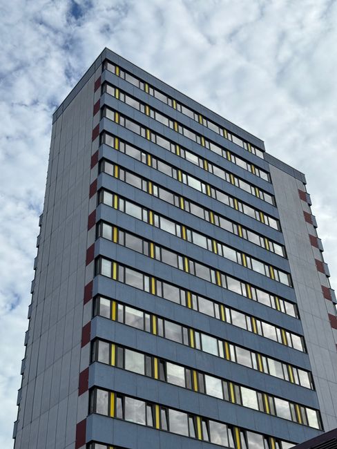 View of a tall modern residential building with multiple floors, featuring large rectangular windows with yellow-tinted glass, set against a partly cloudy sky. The building's facade is predominantly g
