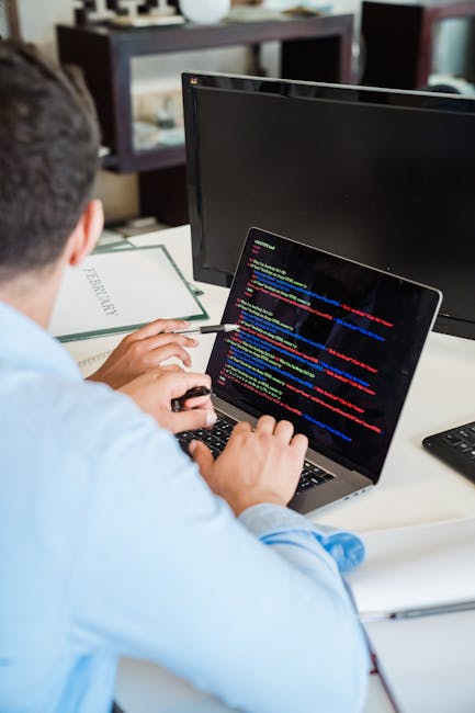 An individual in a light blue shirt seated at a desk engaged in surface cleaning activities related to document or data management, with a laptop displaying colorful code or data on the screen. The wo